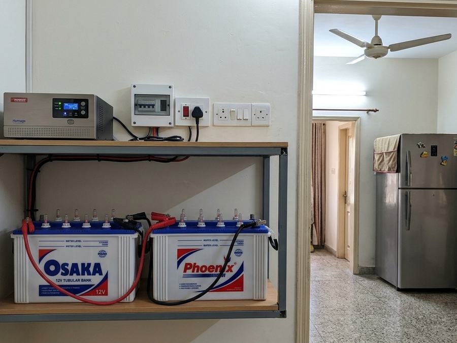 UPS and two batteries on a wall shelf in a home with a fridge and ceiling fan beyond